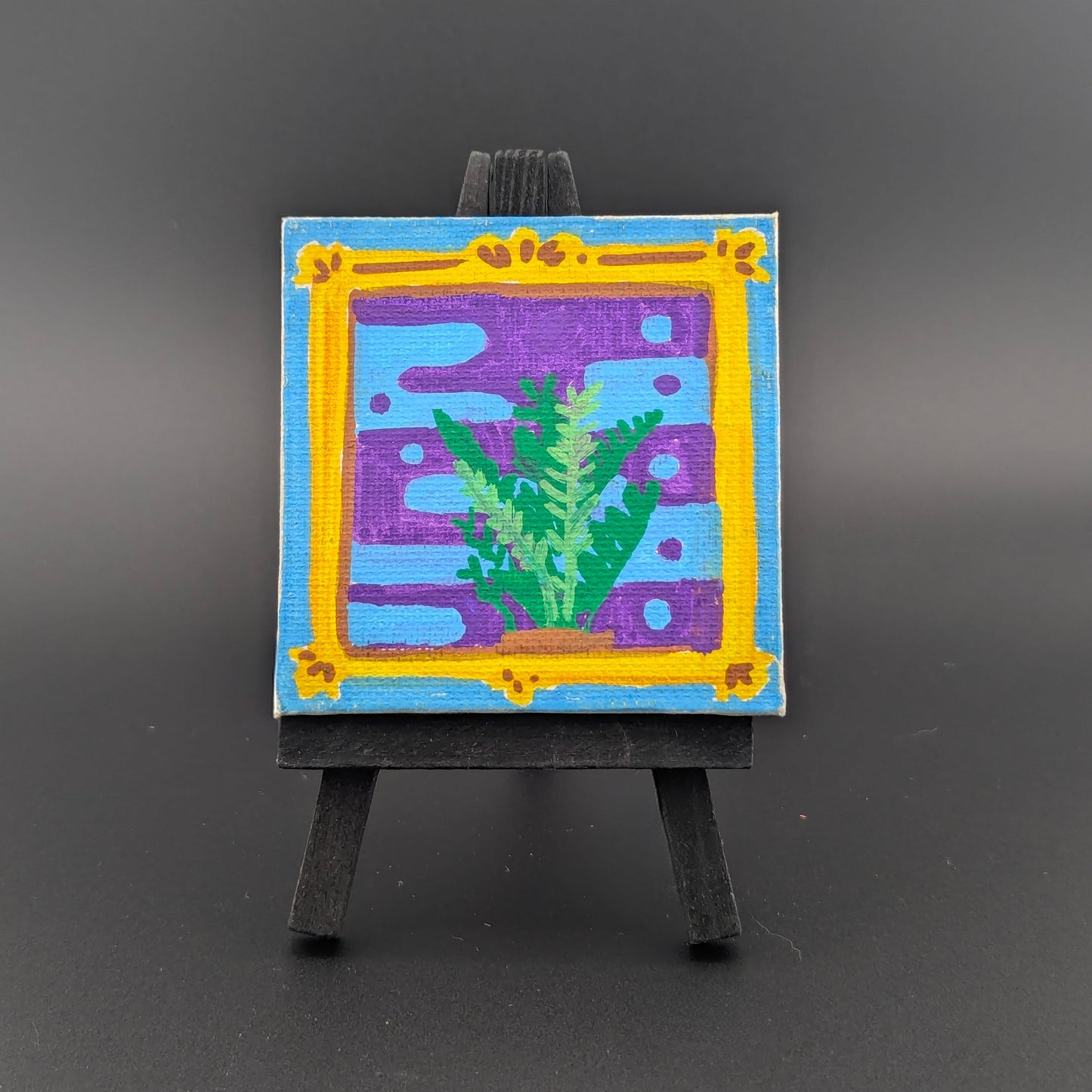 Small painting on an easel with a colorful frame and green plant design on a dark background