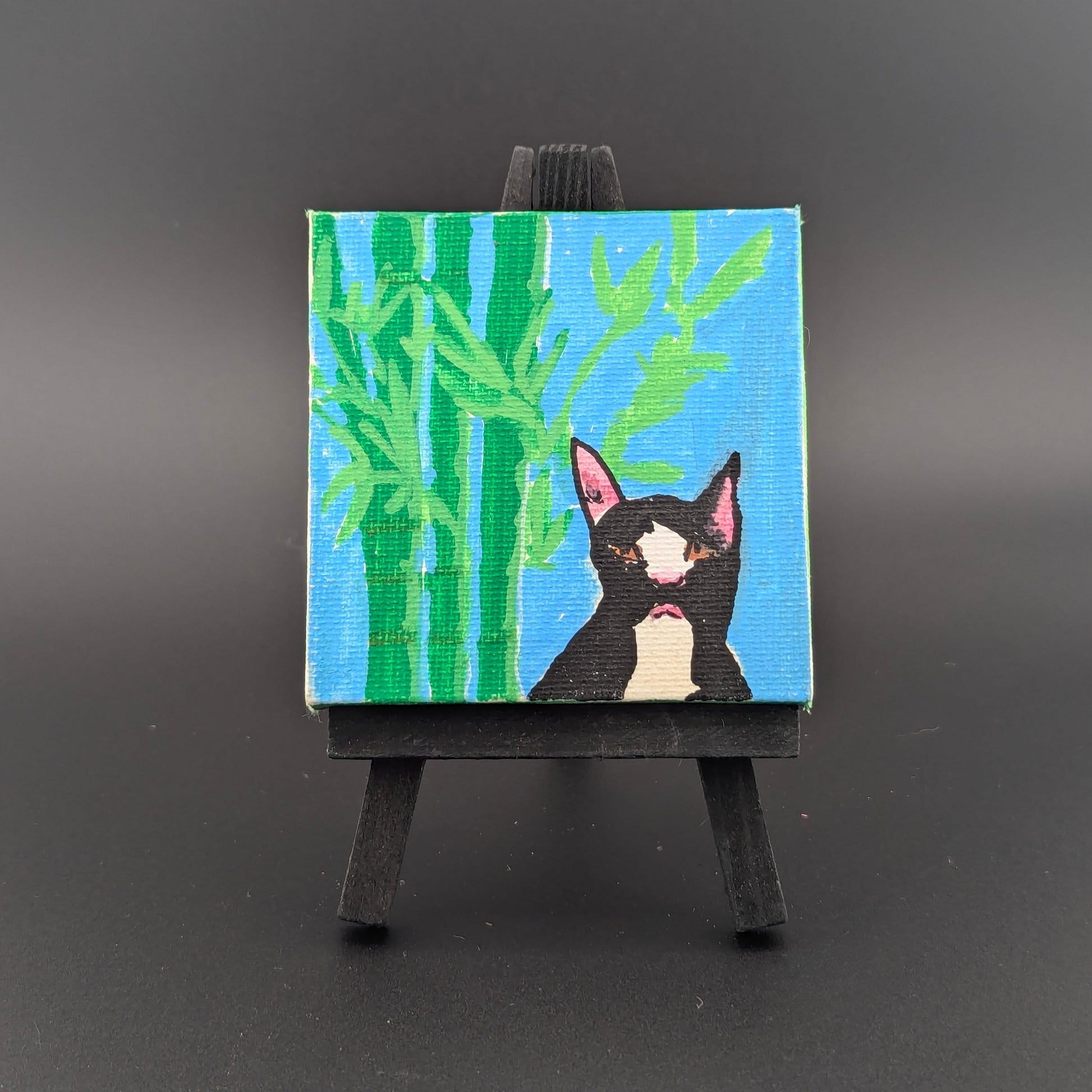 Small painting of a black cat with pink ears on a blue background with green bamboo, displayed on a black easel against a dark gray background.
