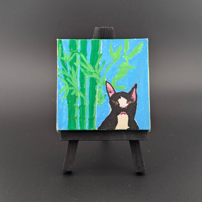 Small painting of a black cat with pink ears on a blue background with green bamboo, displayed on a black easel against a dark gray background.