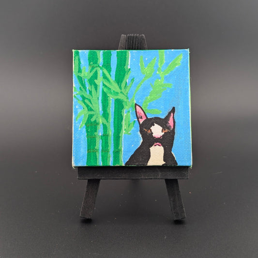Small painting of a black cat with pink ears on a blue background with green bamboo, displayed on a black easel against a dark gray background.